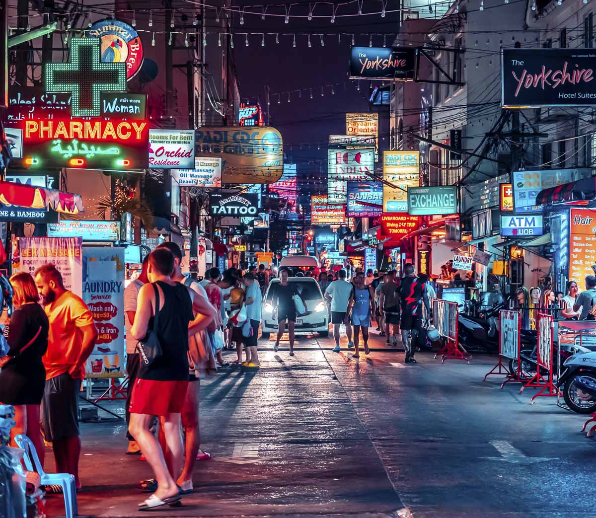 Rua Bangla Road em Phuket à noite, iluminada por letreiros neon de bares, farmácias, casas de câmbio e salões de massagem, com turistas caminhando e carros passando entre a multidão.