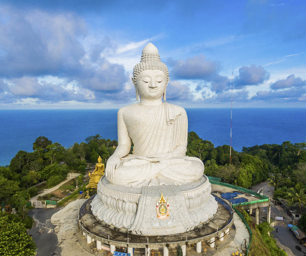 Grande Buda branco de mármore sentado no topo de uma colina em Phuket, com o mar azul e o céu parcialmente nublado ao fundo.
