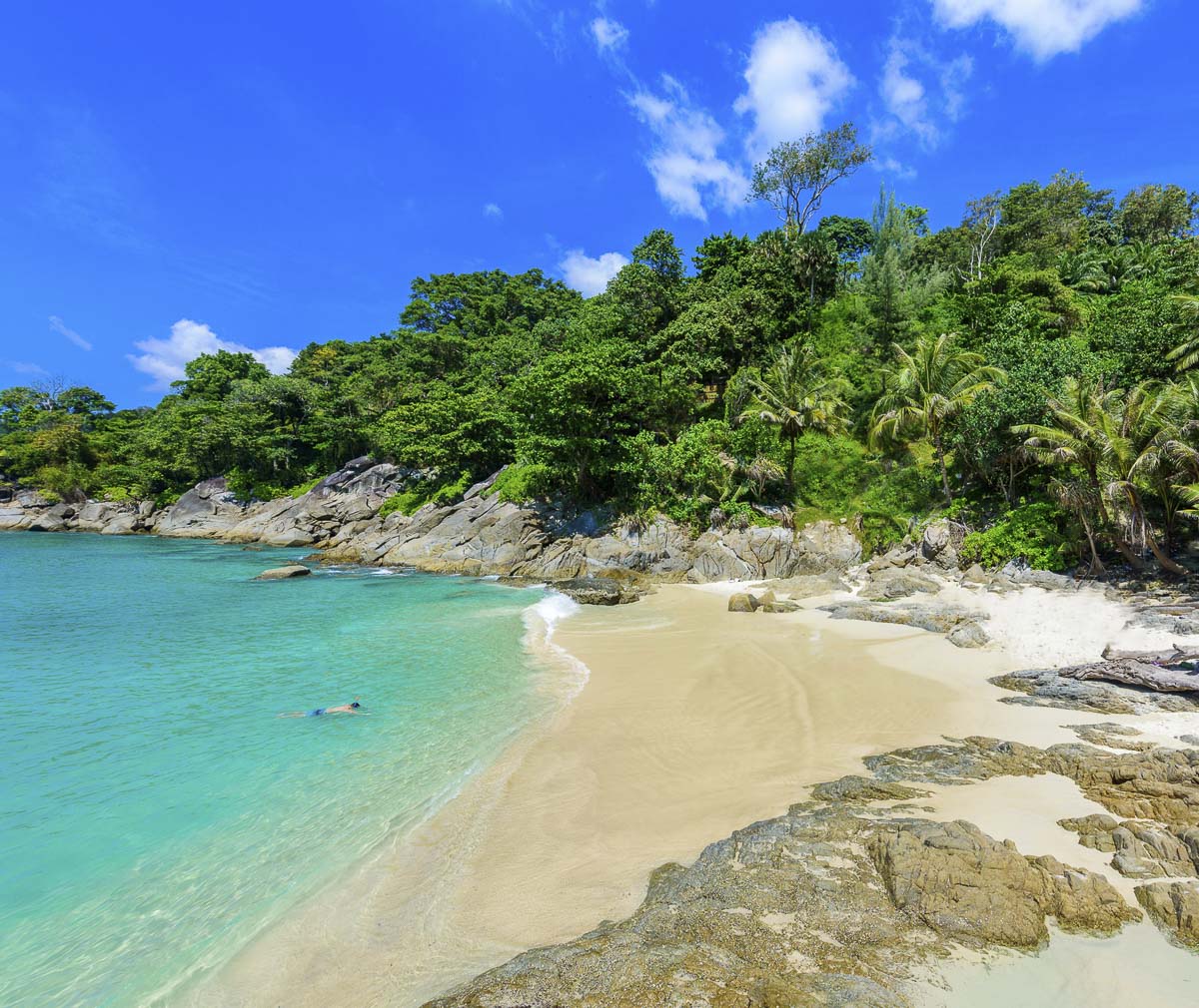 Praia tranquila de areia branca e mar azul-turquesa rodeada por árvores tropicais e rochas, com uma pessoa nadando ao fundo.