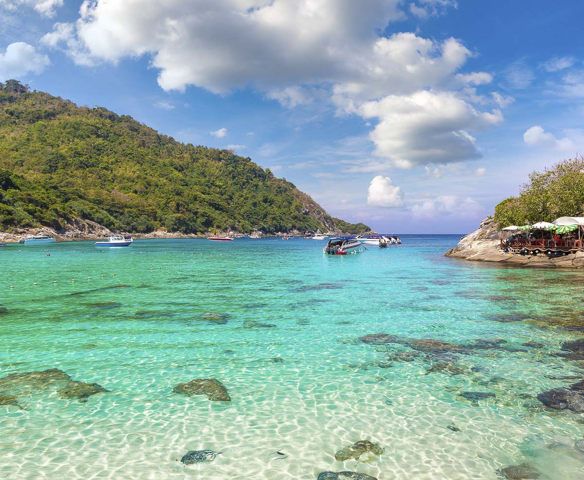 Mar cristalino com tons de azul e verde, barcos ancorados e colinas cobertas de vegetação ao fundo, em Ko Racha Yai, Phuket.