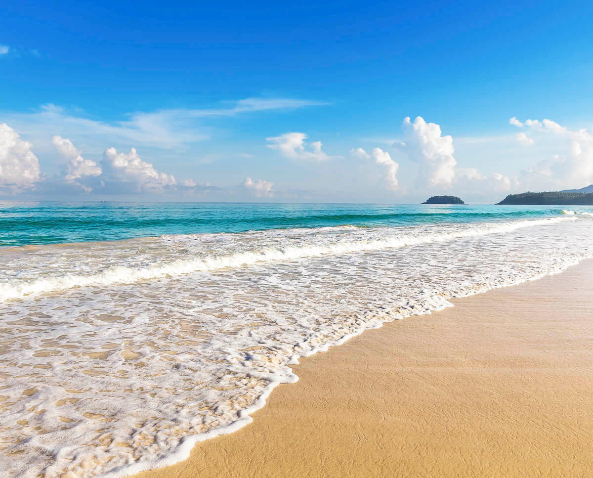 Praia de areia dourada com mar azul e ondas suaves quebrando na areia, sob céu claro com nuvens.