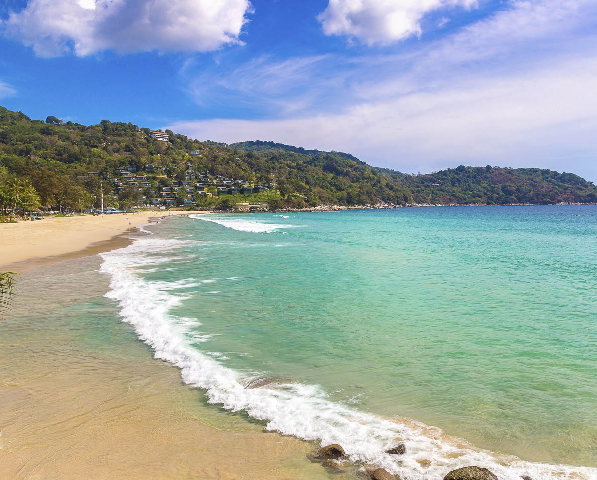 Praia de Kata Noi com mar turquesa, ondas suaves e morros verdes ao fundo sob céu azul com nuvens.