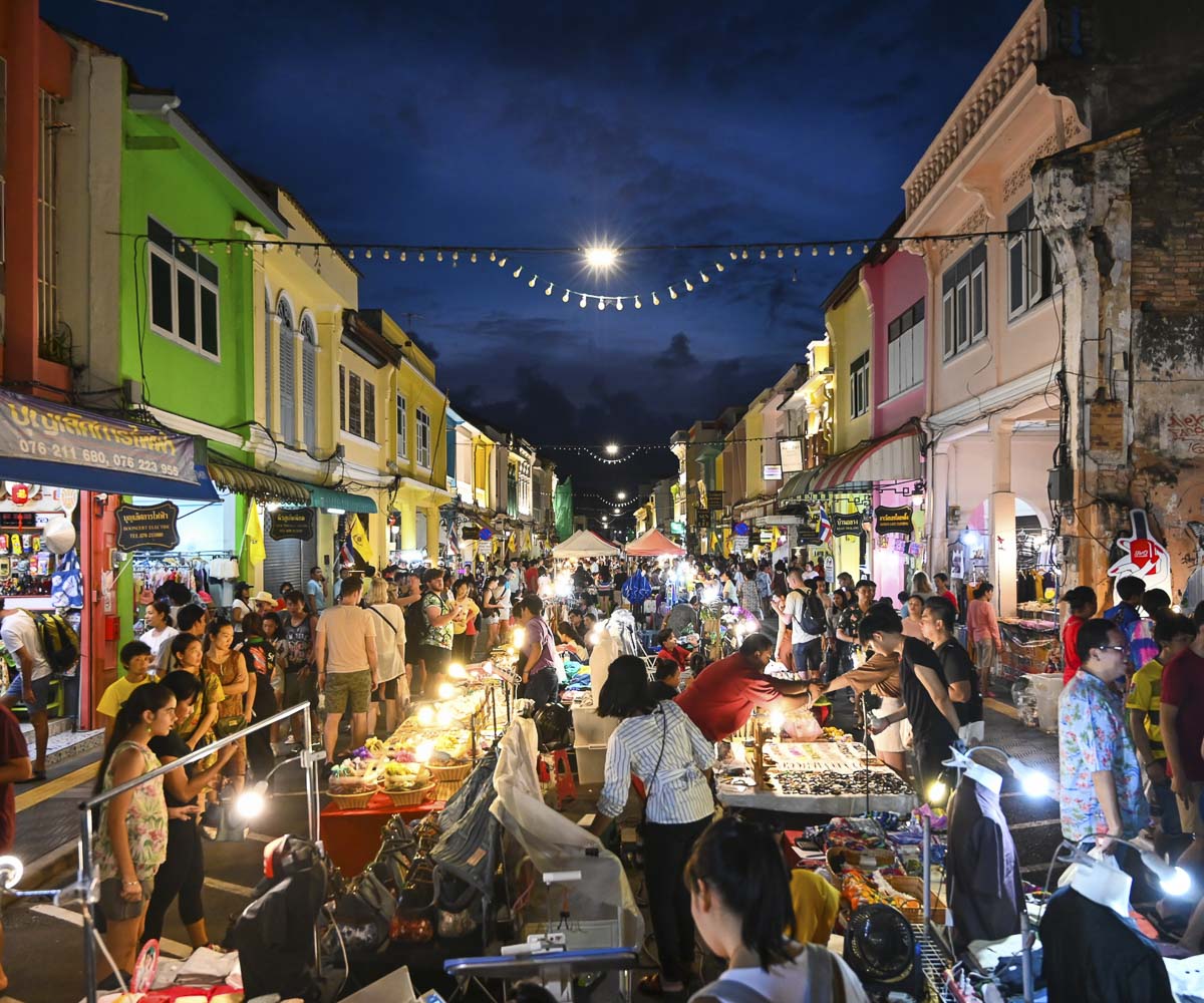 Rua cheia de barraquinhas iluminadas no mercado noturno de Phuket, com multidão entre casas coloridas.