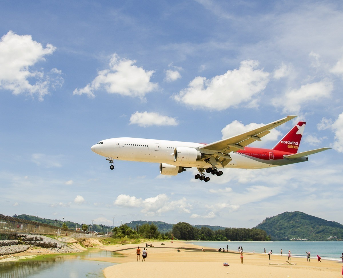 Avião da Nordwind Airlines voa baixo sobre a areia da Mai Khao Beach em Phuket, com turistas observando.