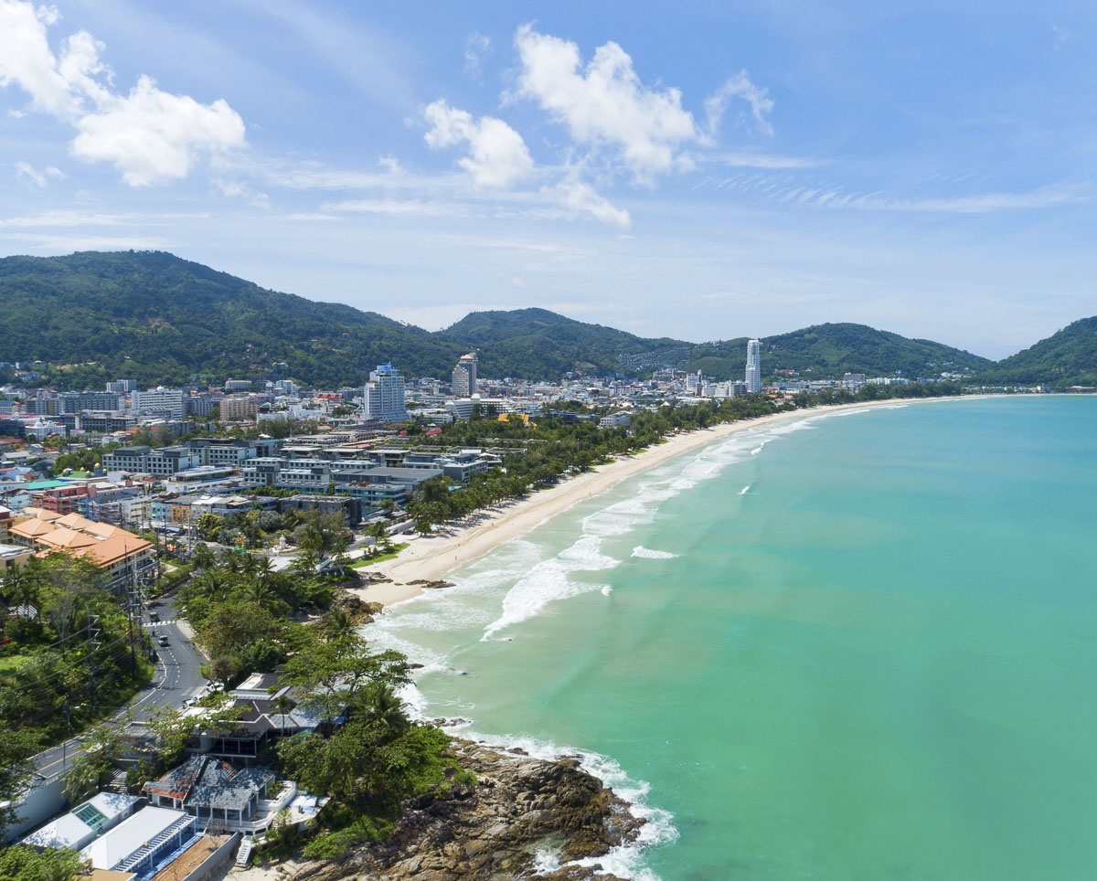 Vista panorâmica da cidade de Patong em Phuket com praia extensa, mar esverdeado e montanhas ao fundo.