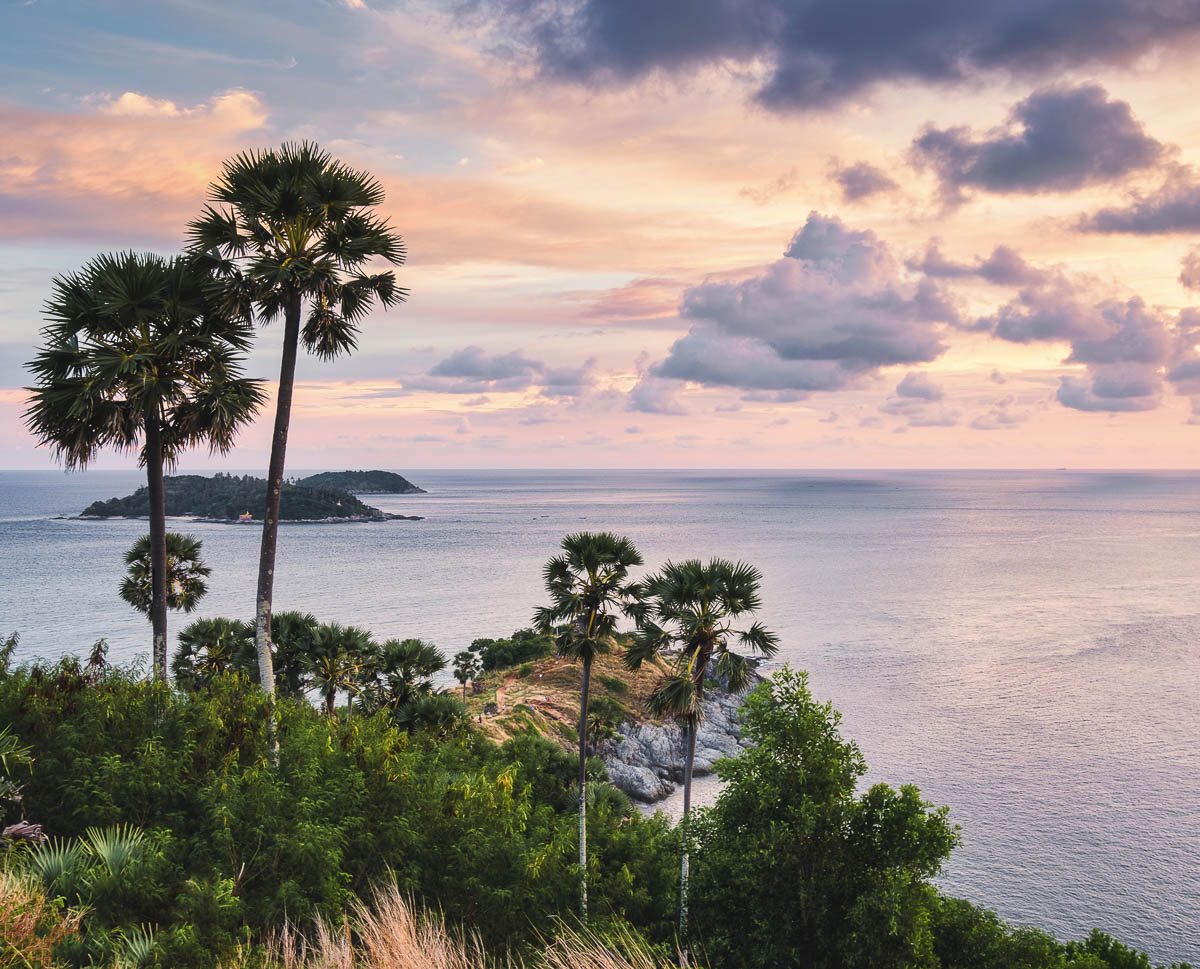 Vista do mar em tons rosa e lilás ao entardecer, com coqueiros altos e uma ilha ao fundo no Promthep Cape.