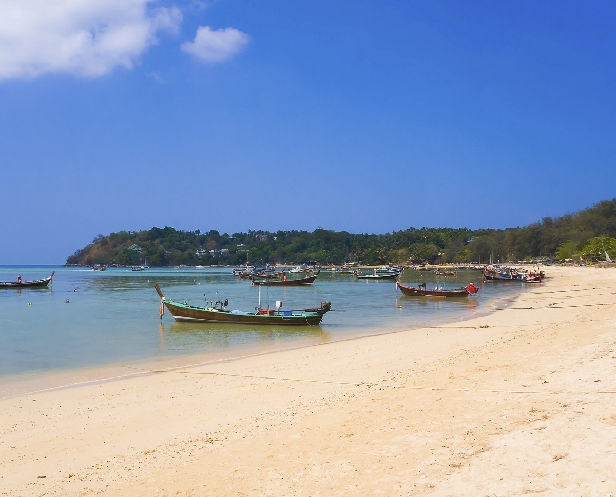 Vários barcos tradicionais longtail ancorados na praia de Rawai, com mar calmo e céu azul.