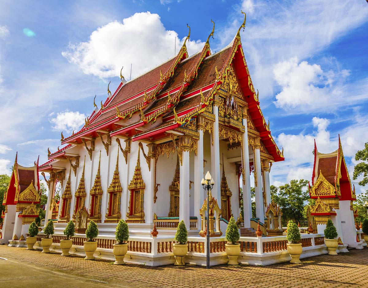 Detalhes dourados e telhados vermelhos do templo Wat Chalong, em Phuket, sob céu azul com nuvens esparsas.