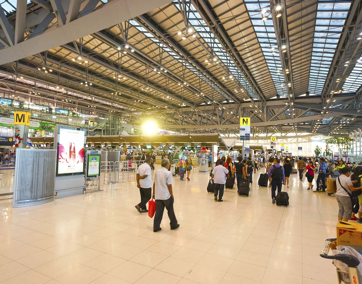 Passageiros com malas caminham em um saguão amplo e iluminado do Aeroporto Internacional de Bangkok, com estruturas metálicas e placas de orientação.