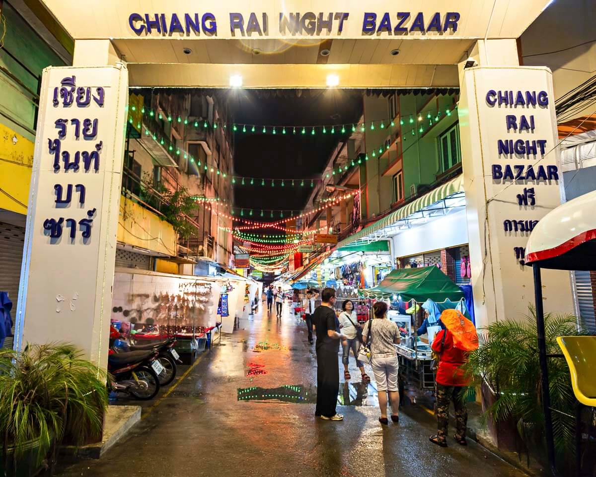 Entrada do Chiang Rai Night Bazaar à noite, com letreiros em tailandês e inglês e luzes coloridas penduradas.