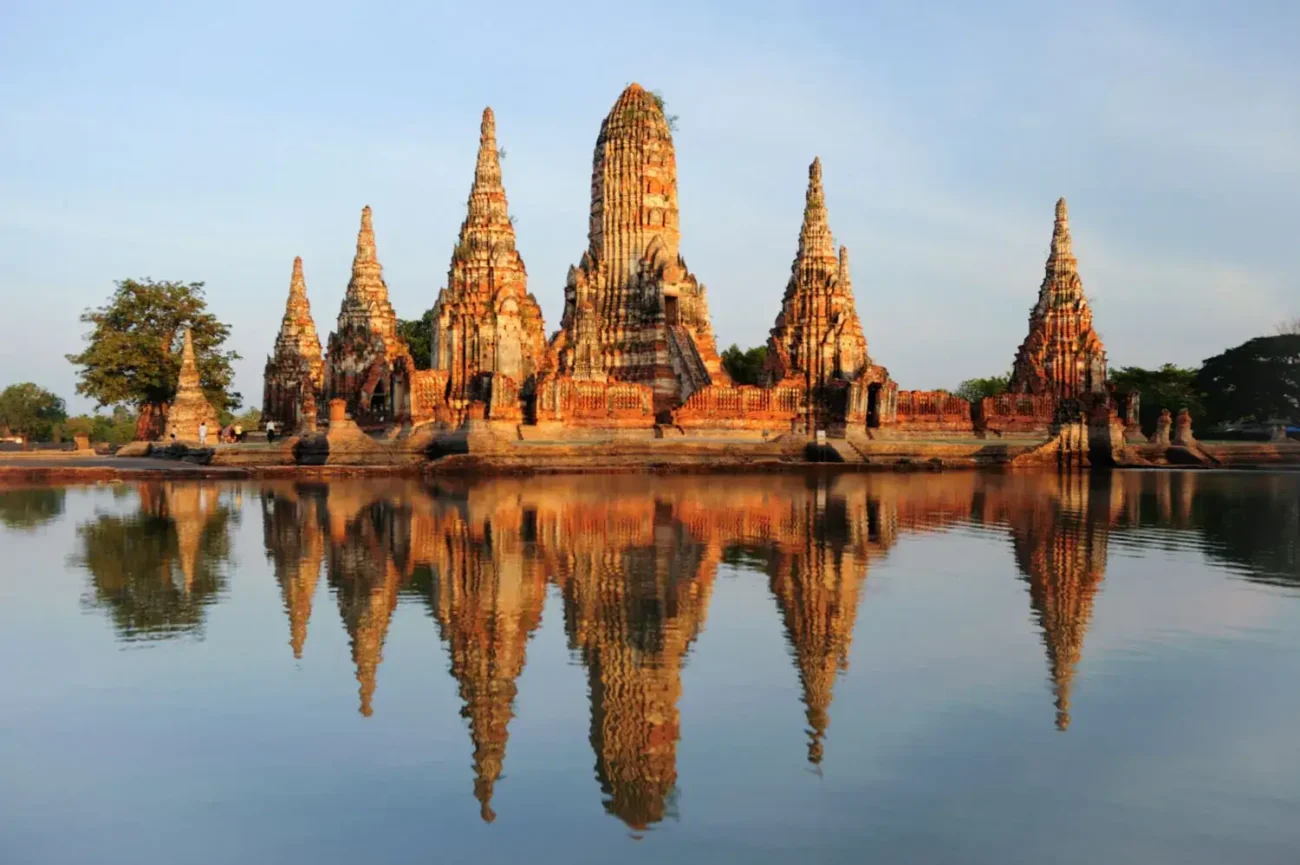 Templo Wat Chaiwatthanaram refletido no rio durante o pôr do sol, com céu limpo ao fundo.