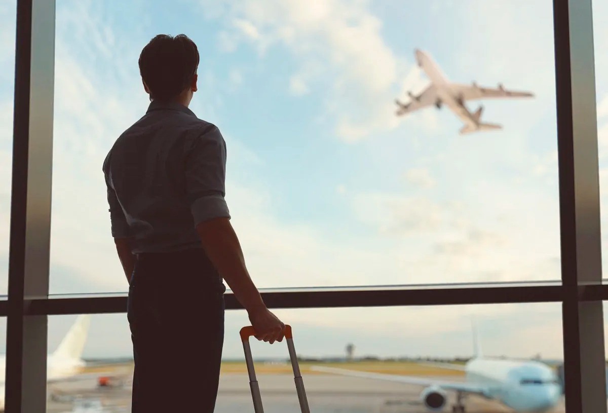 Homem observa avião decolar pela janela do aeroporto, com mala de bordo ao lado.