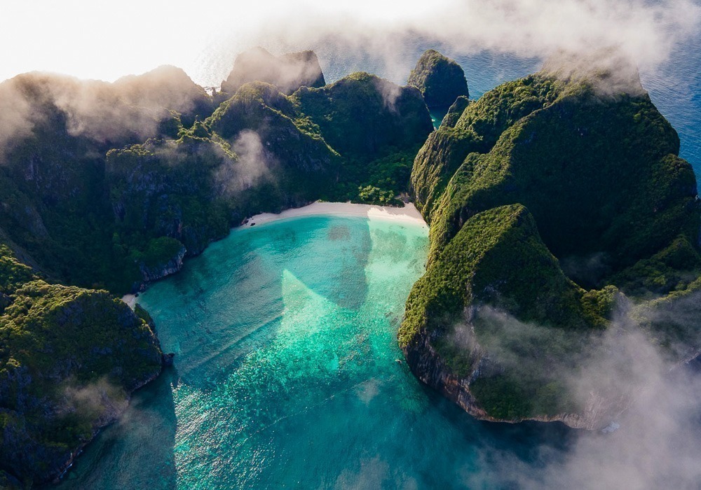 Vista aérea da Maya Bay cercada por falésias verdes e mar turquesa, parcialmente coberta por nuvens.