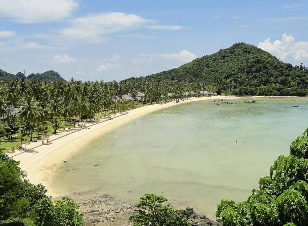 Praia de Lo Ba Kao Bay cercada por coqueiros e vegetação densa, com montanha verde ao fundo.