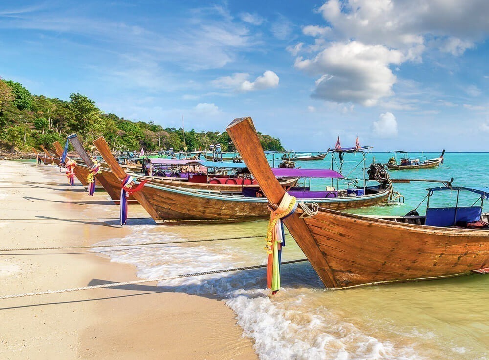 Barcos longtail coloridos ancorados na praia com mar azul e céu parcialmente nublado.