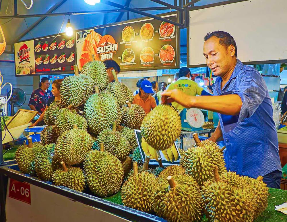 Vendedor manuseando frutas durian em uma barraca colorida no mercado noturno de Ao Nang, em Krabi.