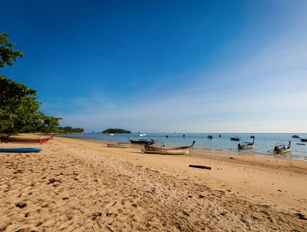 Barcos longtail ancorados em uma praia tranquila com areia dourada e céu azul em Klong Muang, Krabi.
