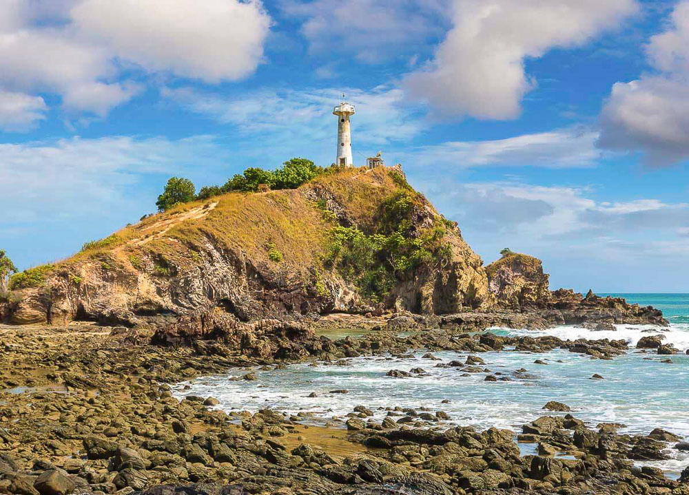 Farol branco sobre colina rochosa com vegetação rala, rodeado por mar e céu parcialmente nublado em Ko Lanta.