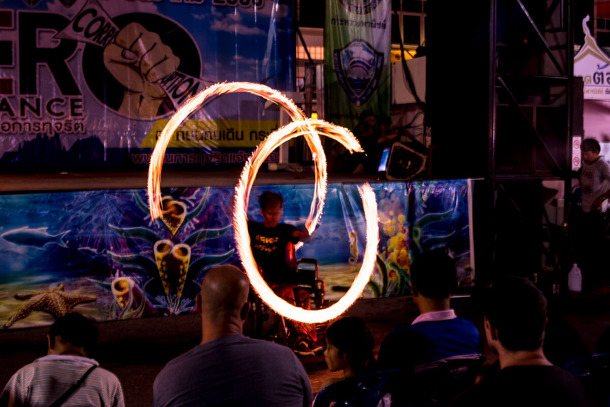 Artista em cadeira de rodas se apresenta com malabares de fogo em palco no mercado noturno de Krabi Town.