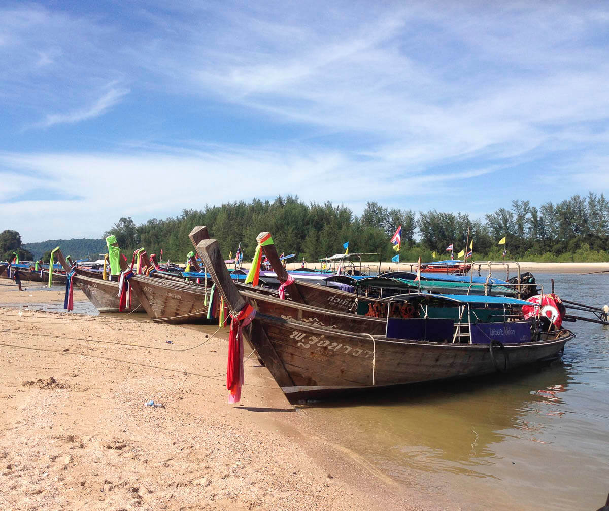 Barcos longtail tradicionais tailandeses ancorados na areia da tranquila Nopparat Thara Beach, com céu azul e vegetação ao fundo.