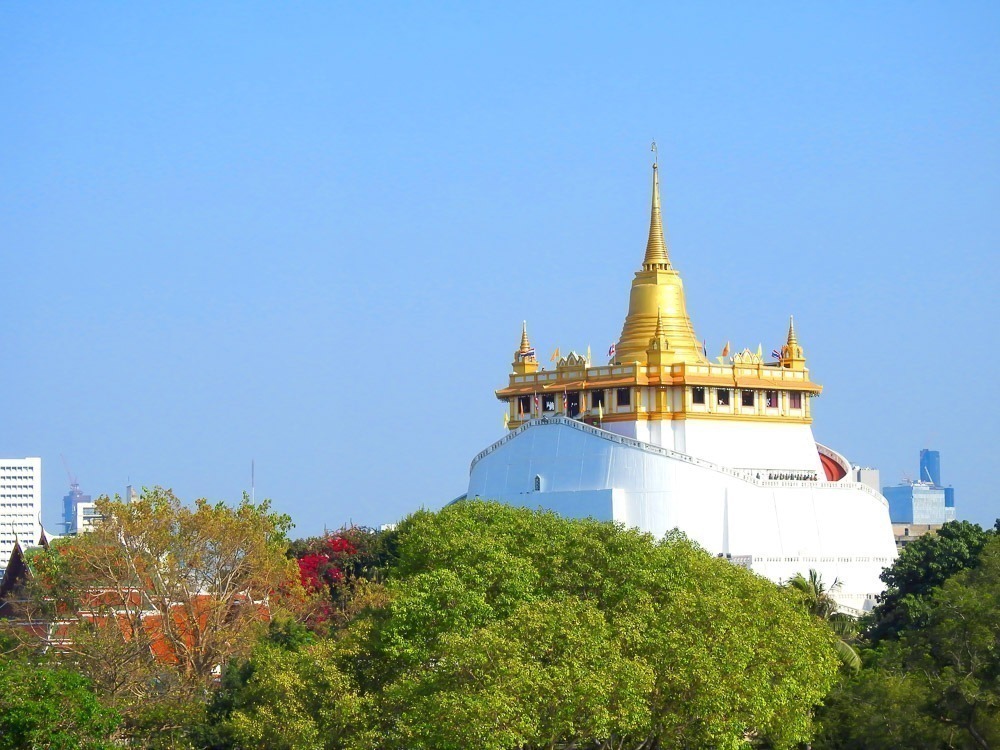 Templo Wat Saket com sua estupa dourada no topo de uma colina branca cercada por vegetação e prédios urbanos ao fundo.