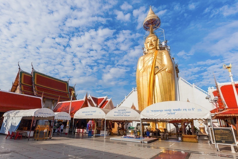 Grande estátua dourada de Buda em pé no Wat Intharawihan, rodeada por tendas brancas e visitantes.