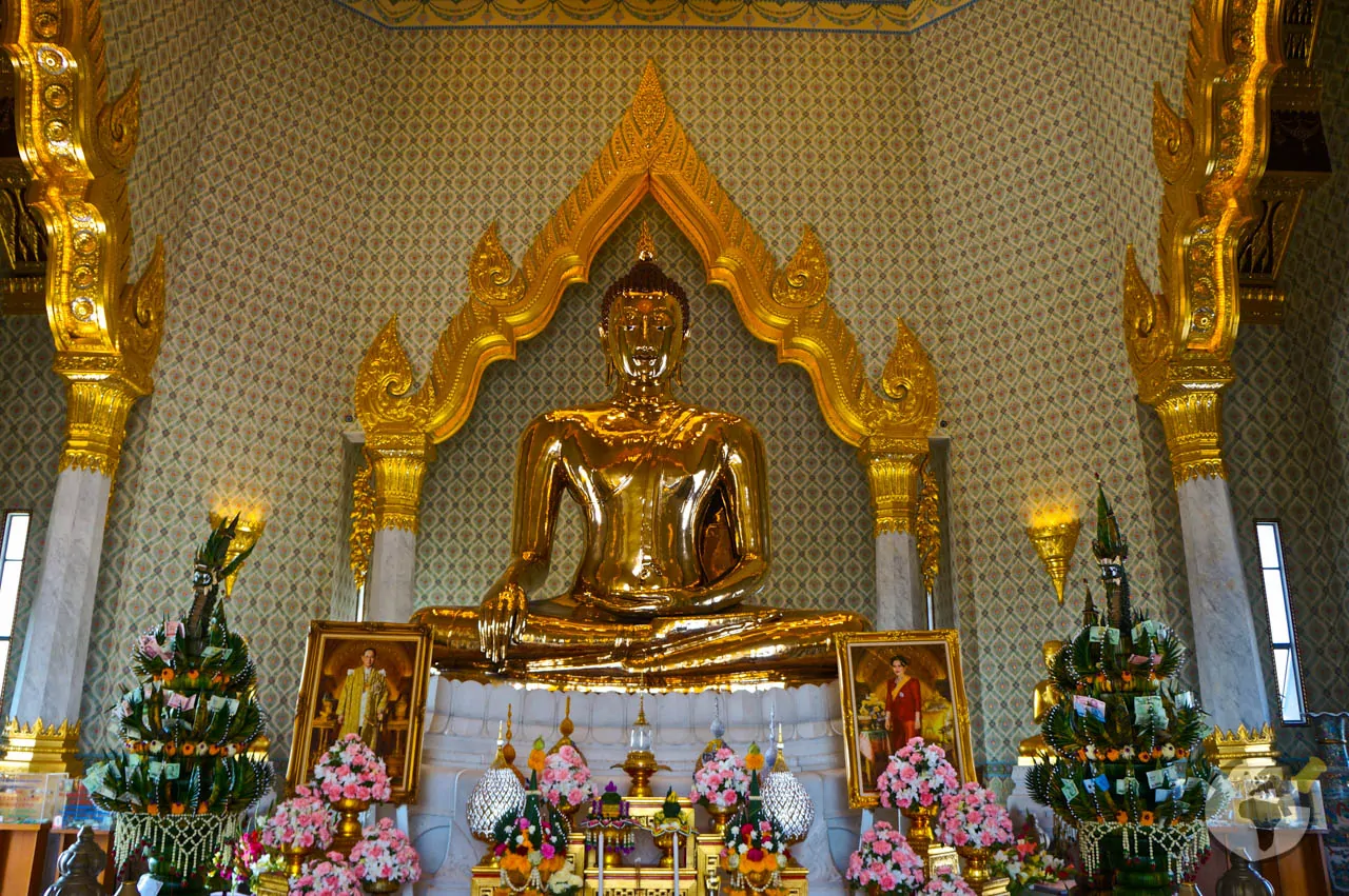Estátua dourada do Buda de Ouro no Wat Traimit, cercada por flores e quadros dentro do templo.