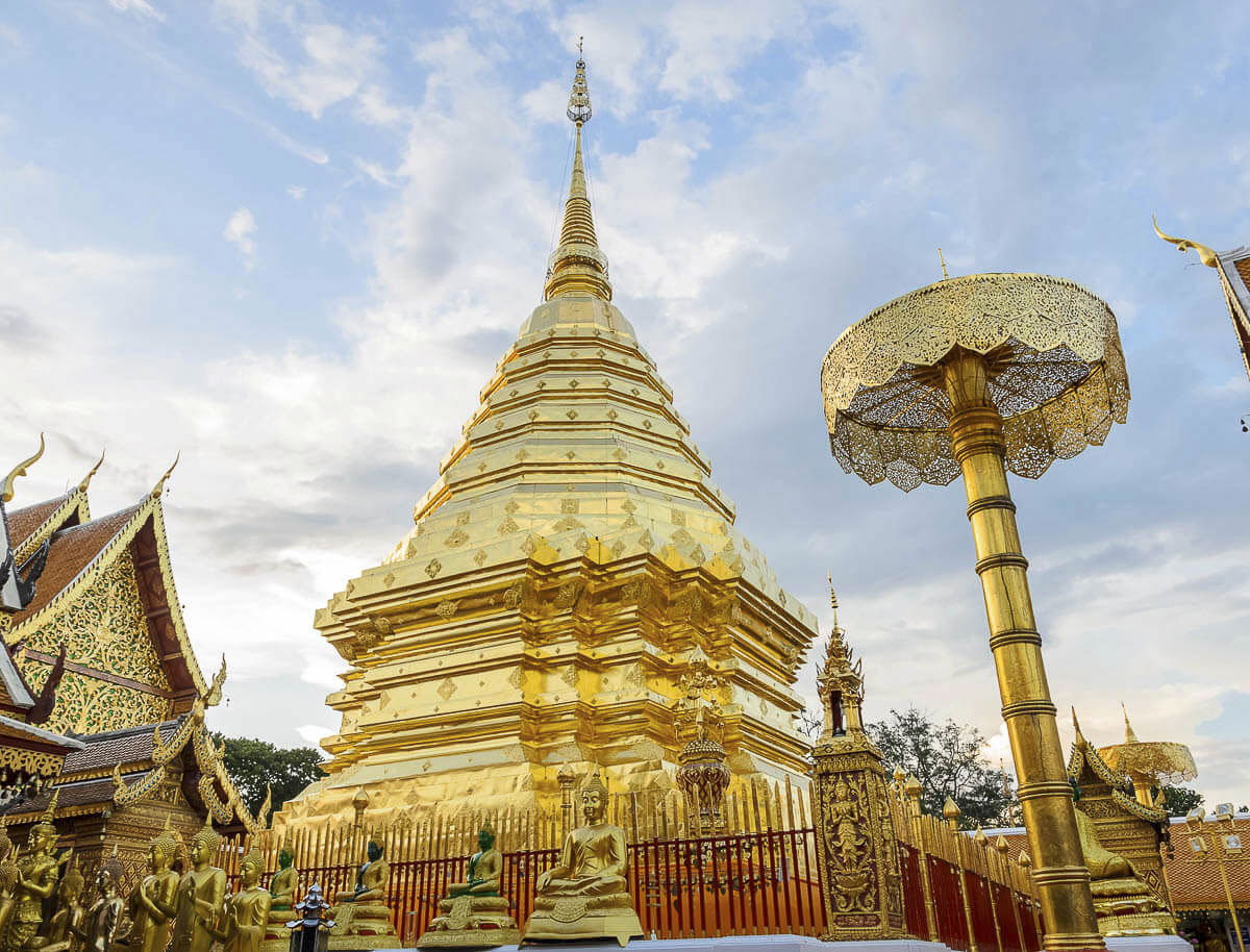 Chedi dourado do Wat Phra That Doi Suthep em Chiang Mai, com esculturas budistas ao redor e céu nublado ao fundo.