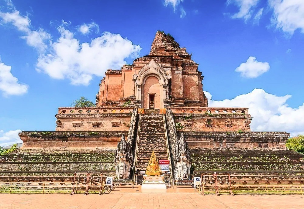 Escadaria frontal e estrutura antiga em tijolos do Wat Chedi Luang, com céu azul e nuvens ao fundo.