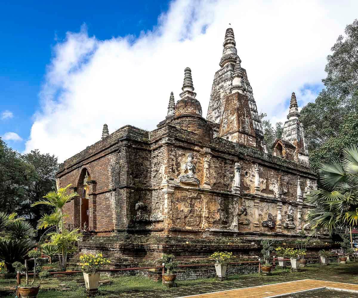 Estrutura retangular de tijolos antigos com sete torres pontiagudas do Wat Chet Yot, cercada por vegetação.