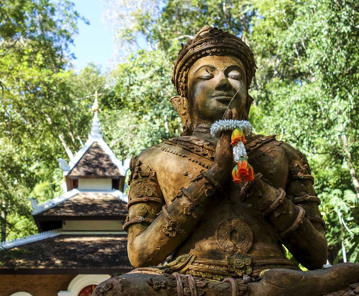 Estátua de divindade budista em oração, com guirlanda de flores e fundo de floresta no Wat Pha Lat.