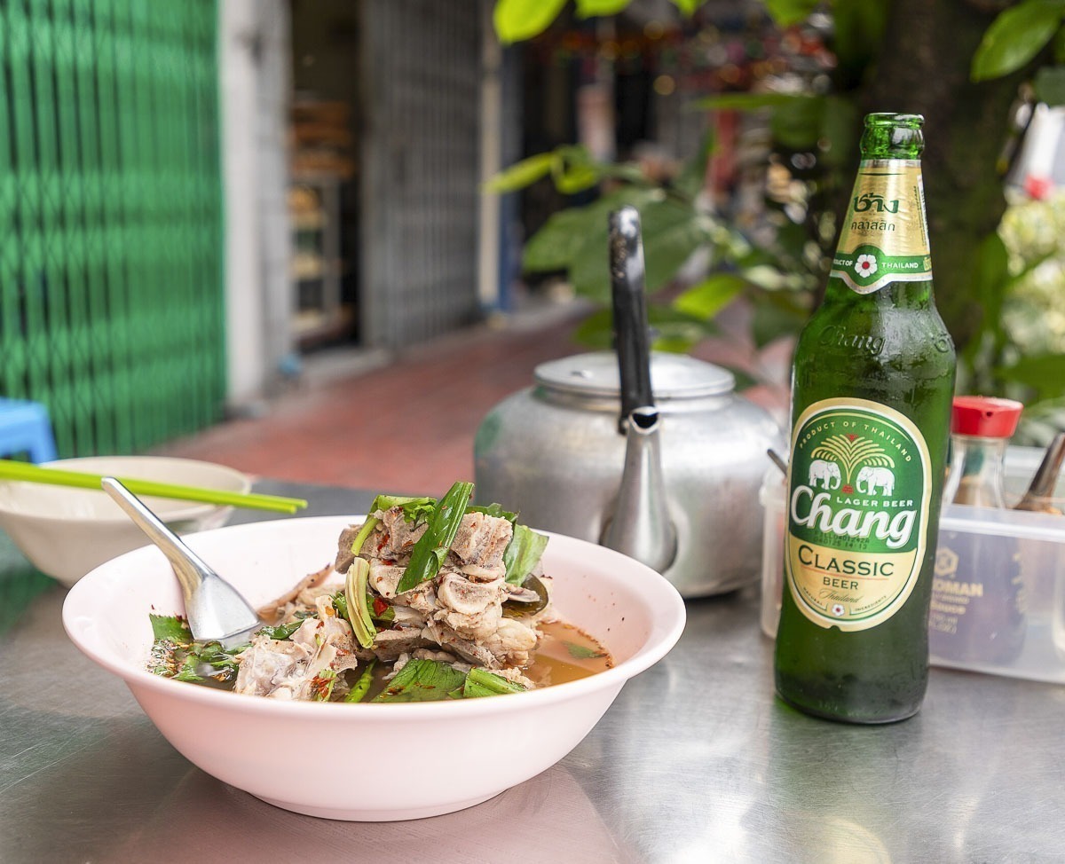 Tigela de sopa tailandesa ao lado de uma garrafa de cerveja Chang sobre mesa de rua.