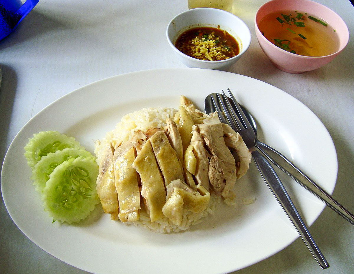 Prato de khao man gai com fatias de frango, arroz e pepino, acompanhado de molho e sopa.