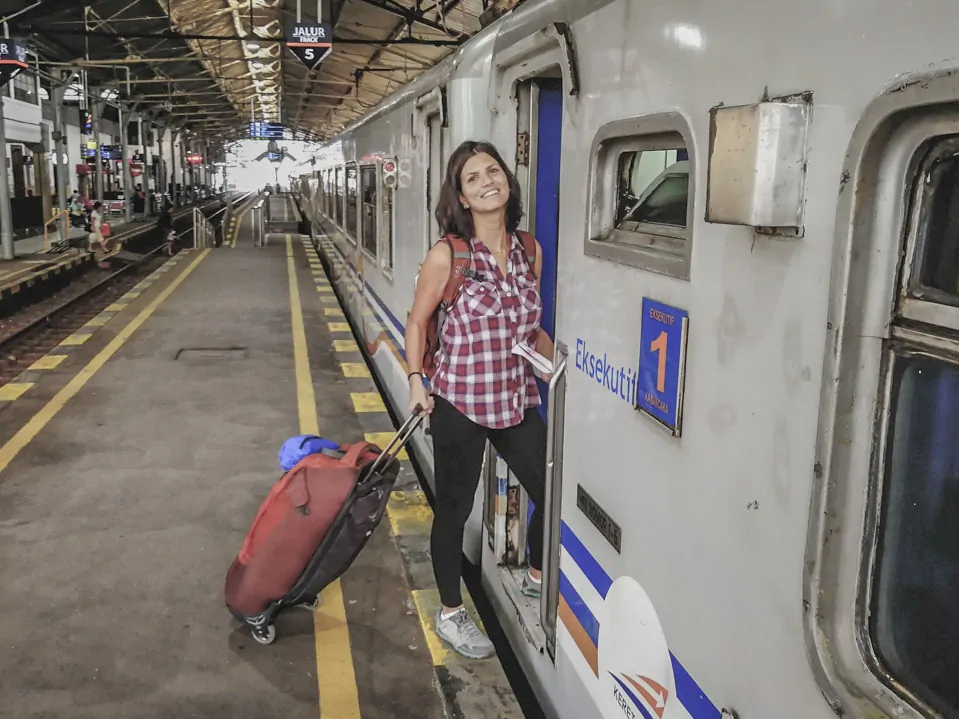 A Dri prestes a embarcar em um trem na estação, com mochila nas costas e mala de rodinhas ao lado.