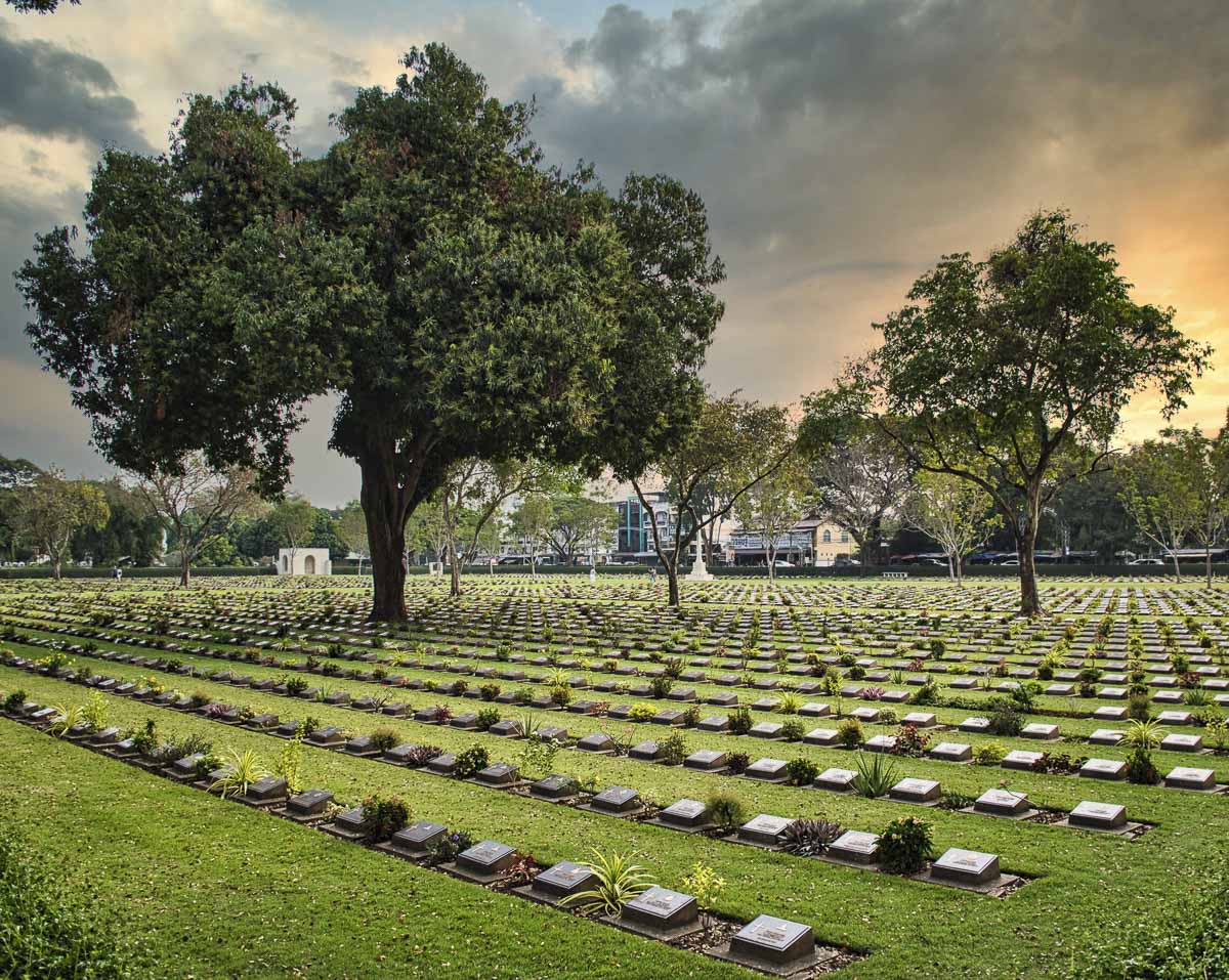 Cemitério de guerra em Kanchanaburi com lápides alinhadas e grandes árvores ao pôr do sol.