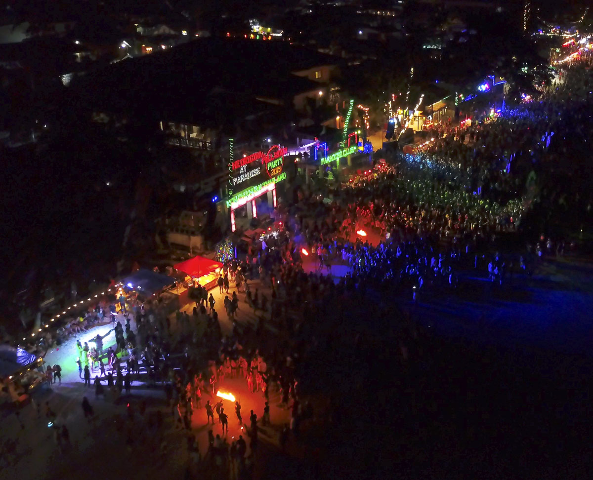 Festa noturna em Koh Phangan com milhares de pessoas reunidas, luzes neon e apresentação com fogo na areia.