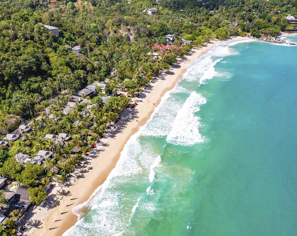 Praia com mar azul vibrante e resorts entre coqueiros em Koh Phangan.