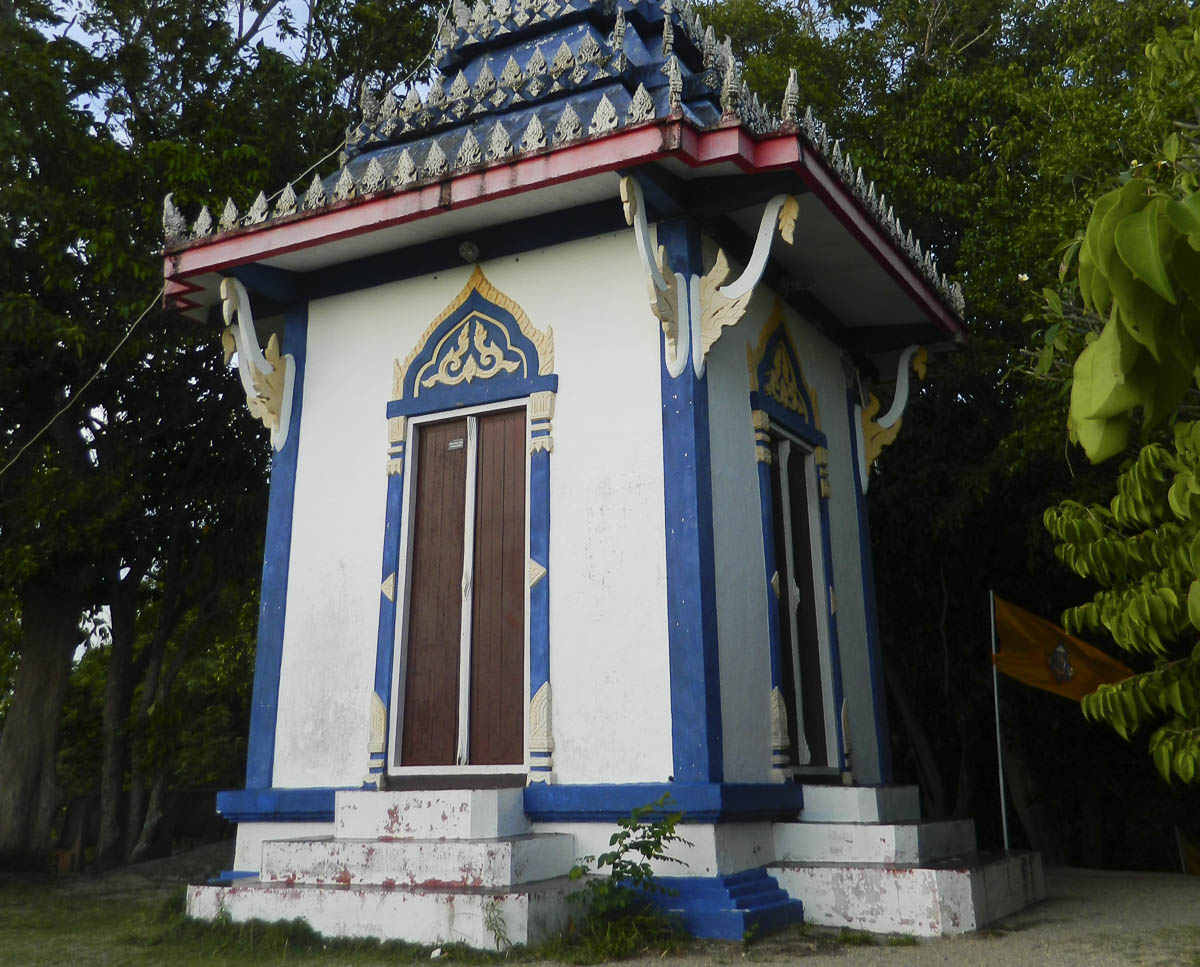 Pequeno templo em estilo tailandês, com detalhes azuis e dourados, cercado por árvores.