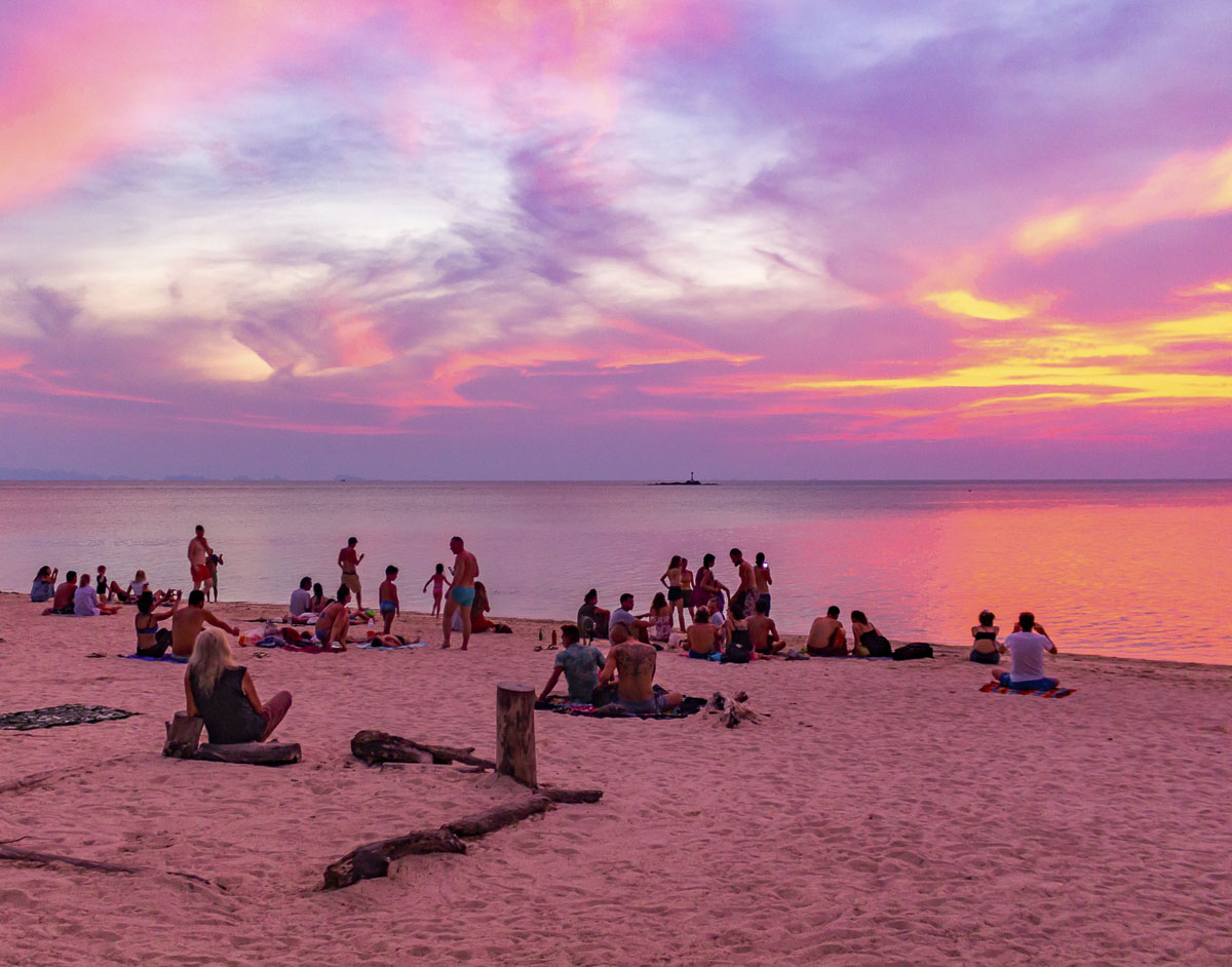 Pessoas reunidas na areia de Koh Phangan observando o pôr do sol com céu rosa e roxo.