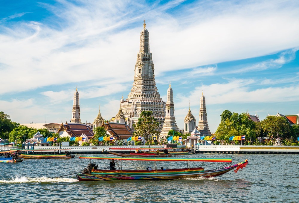Templo Wat Arun com torres brancas ornamentadas refletindo à beira do rio Chao Phraya, com barcos coloridos no primeiro plano.