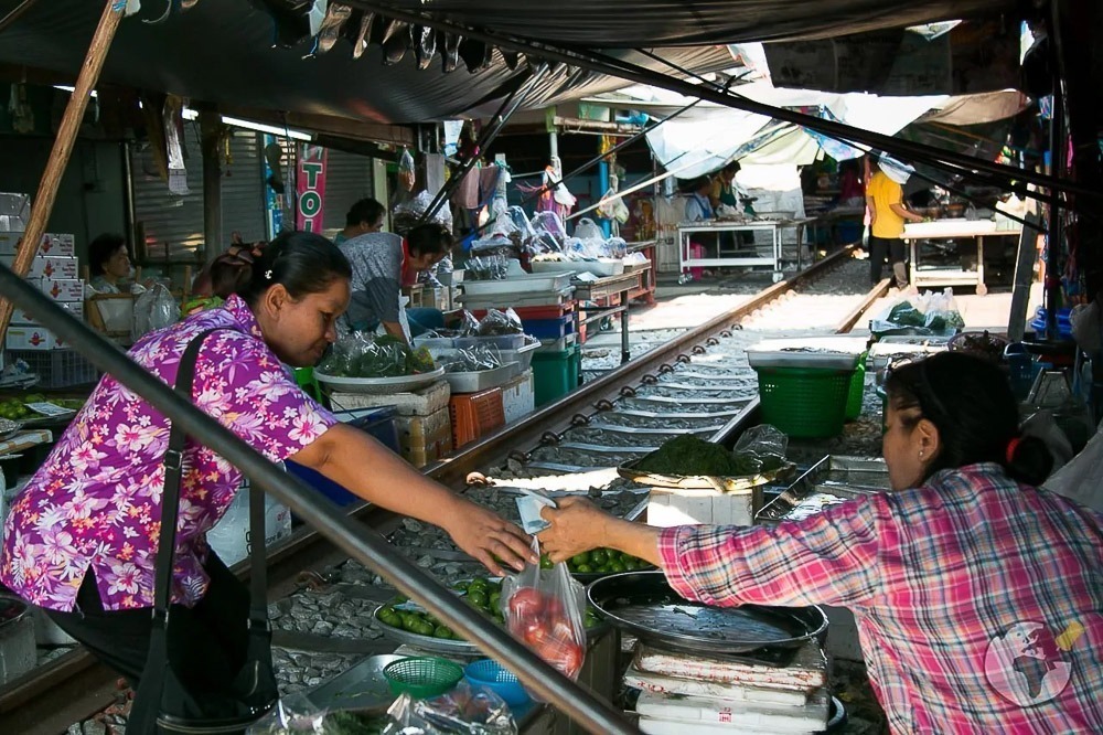 Vendedoras trocando mercadorias em meio às barracas do Maeklong Railway Market, com trilhos passando entre os estandes.