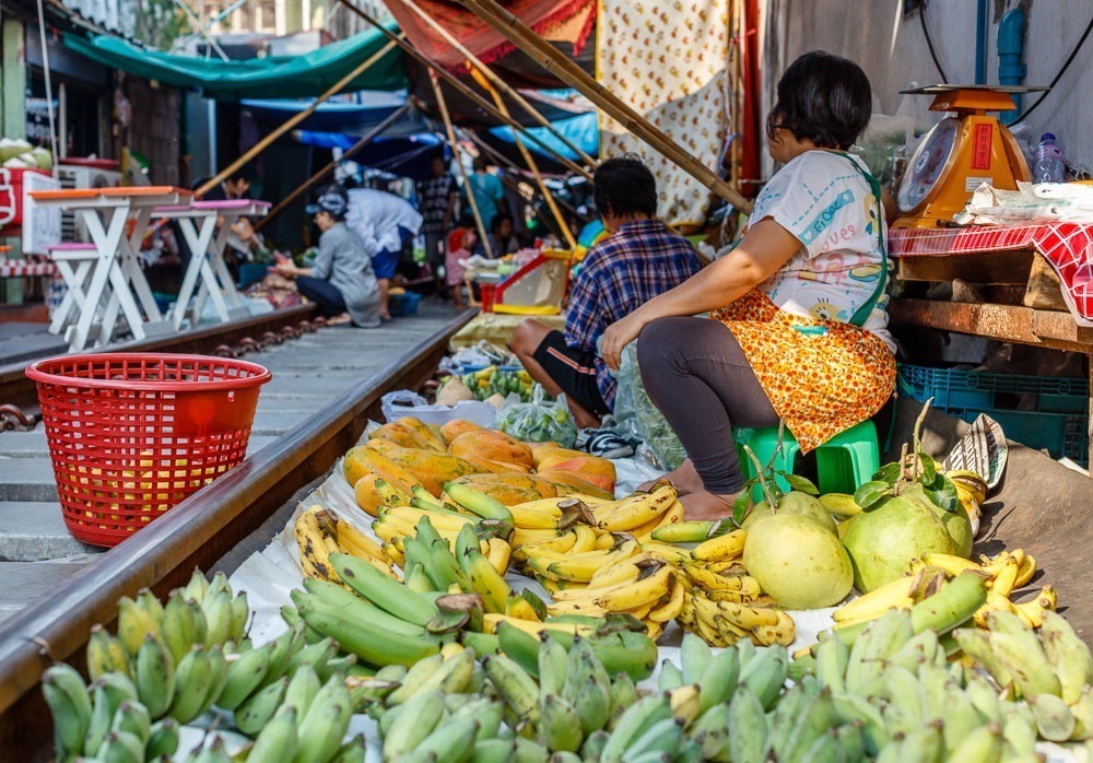 Vendedora sentada ao lado de uma grande variedade de frutas tropicais, incluindo bananas e mamões, no Maeklong Railway Market.
