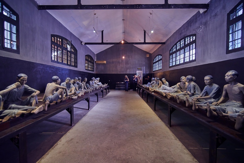 Sala escura da Prisão de Hỏa Lò com esculturas de prisioneiros acorrentados sentados em fileiras.
