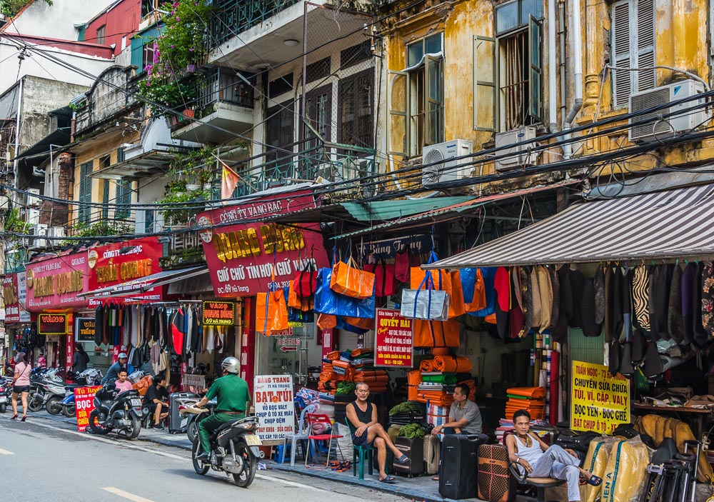 Rua movimentada no bairro antigo de Hanói com lojas, fios expostos e prédios coloniais desgastados.