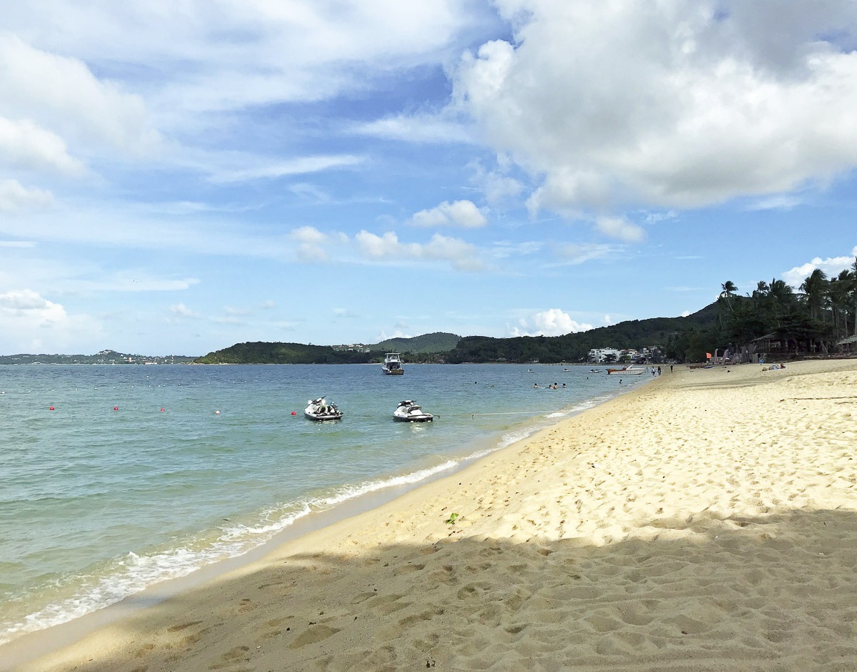 Praia de areia clara em Koh Samui com jet skis ancorados e mar azul tranquilo sob céu parcialmente nublado.