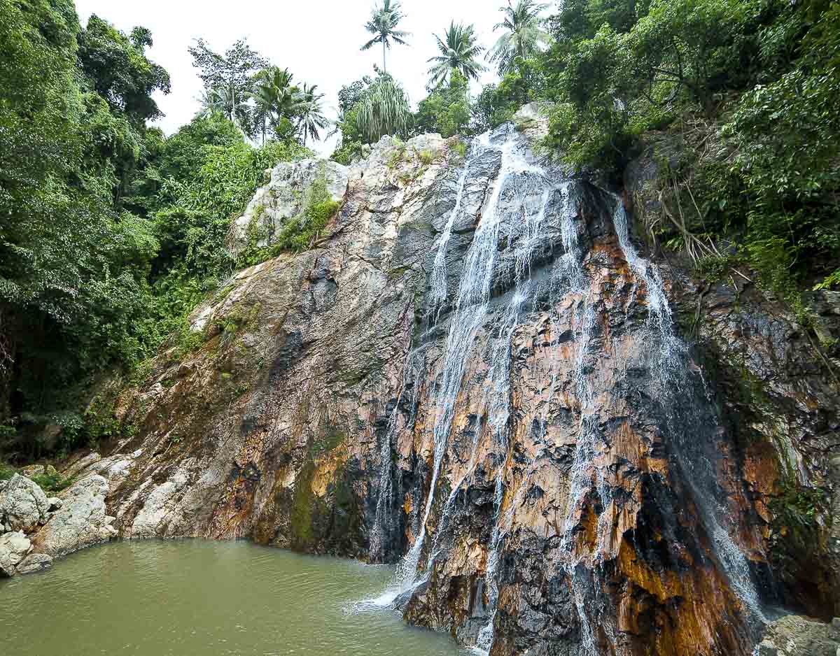 Cachoeira descendo por rochas verticais escuras cercadas por vegetação densa em Koh Samui.