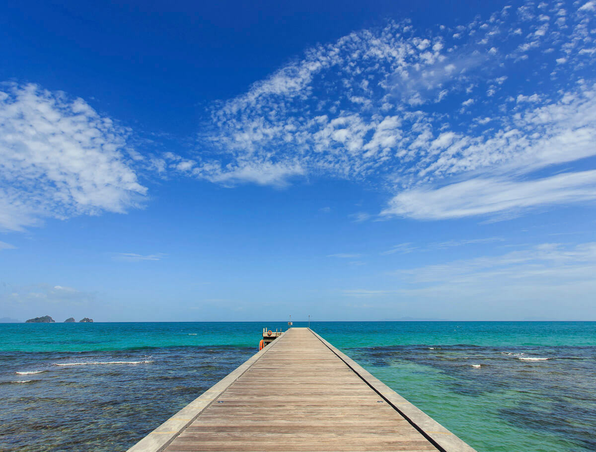 Píer de madeira longo se estende até o mar azul-turquesa sob céu azul com nuvens espalhadas em Koh Samui.