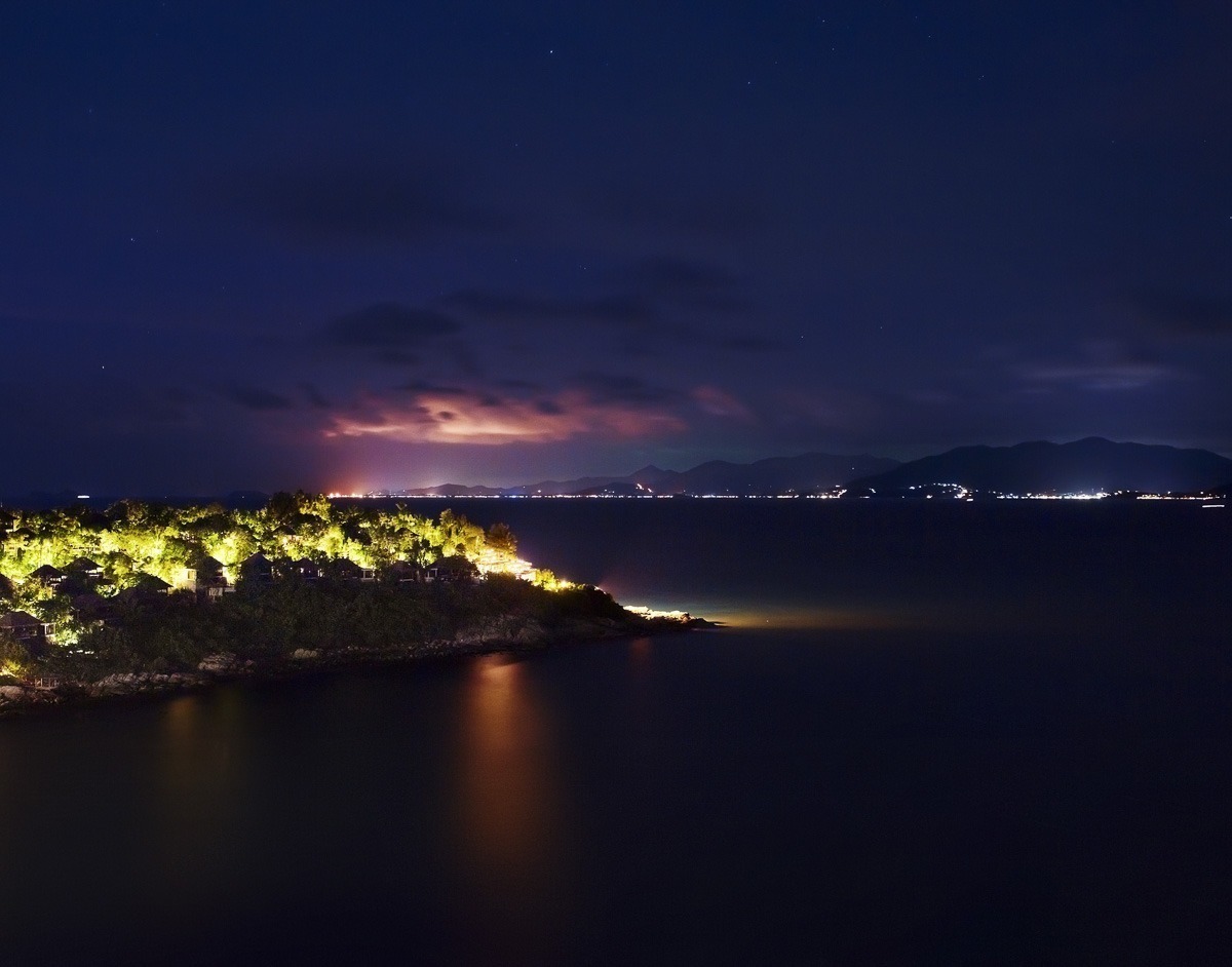 Vista noturna da costa de Koh Samui com luzes nas encostas e céu escuro iluminado por relâmpagos ao fundo.