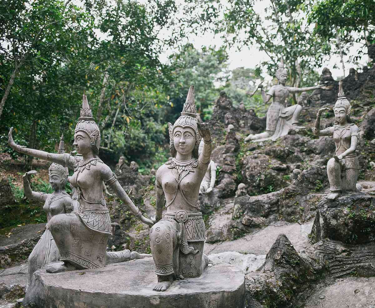 Estátuas de figuras mitológicas e dançarinas tailandesas esculpidas em pedra em meio à vegetação no Tarnim Magic Garden.