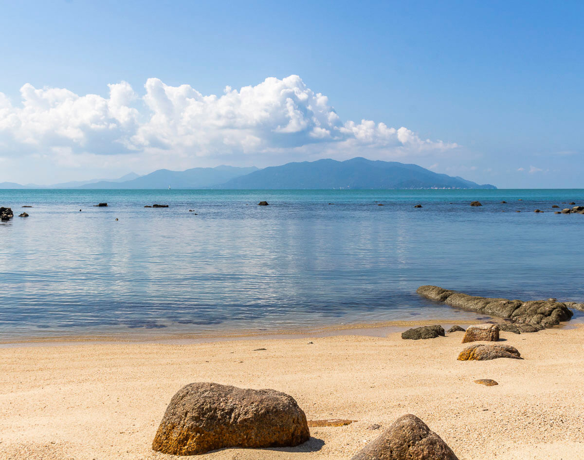 Praia de areia clara com pedras espalhadas e vista para o mar azul calmo com montanhas ao fundo em Koh Samui.